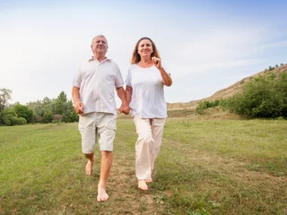 Senior man and senior woman holding hand walking barefoot on green field. Cheerful mature couple enjoying active lifestyle in nature. Concept of healthy retirement and togetherness. © Tatyana Gladskih