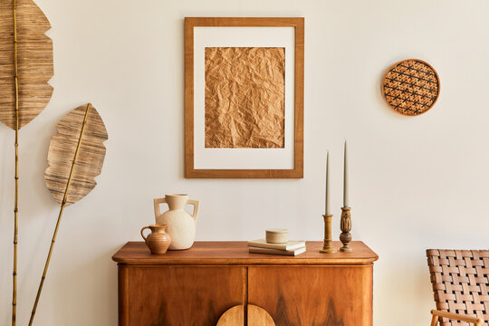 Minimalist composition of living room with brown mock up picture frame, plant, retro armchair, dried tropical leaf, decoration and elegant personal accessories in stylish home decor. Template.