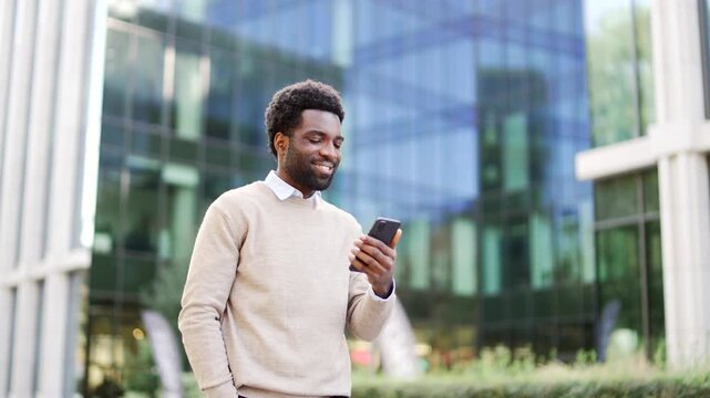 Smiling african american businessman walking on street holding using smartphone on the street outside office building. Confident happy entrepreneur in casual sweater checking mobile phone in urban cit