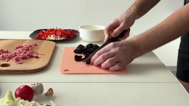 Hands chopping dried prunes on pink cutting board with diced ingredients arranged on white countertop in bright kitchen scene, wooden board, black plate, garlic, tomato and pepper nearby
