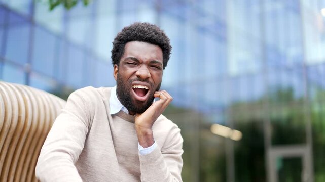African american businessman suffering from toothache outside on street near business office building. Upset man holds his hand on his cheek, massaging sore spot, needs treatment at dentist. Close up