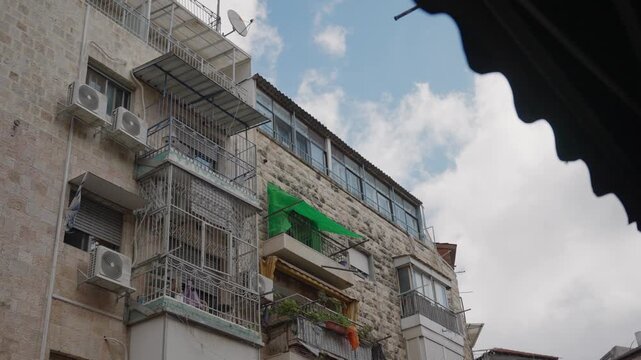 Low angle view of a traditional stone apartment building with balconies and air conditioning units, standing against a blue sky with clouds, filmed from a narrow street in Jerusalem