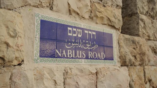 Ornate ceramic street sign for Nablus Road in Jerusalem written in Hebrew, Arabic, and English, mounted on a weathered, ancient stone wall of the old city