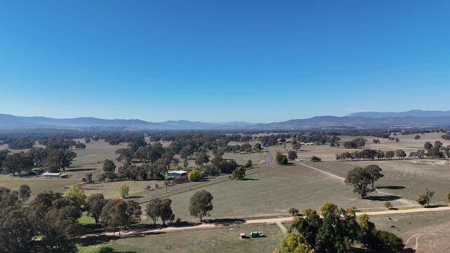 Aerial panoramic view of a vast rural countryside landscape with scattered trees, a winding road, and distant mountains under a clear blue sky.