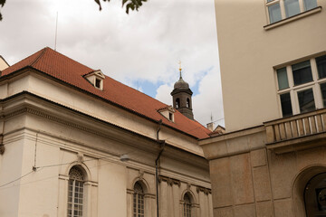 Partial view of a historic church with red tiled roof and arched windows nestled among adjacent urban buildings. A small bell tower with a cross rises above © Alena