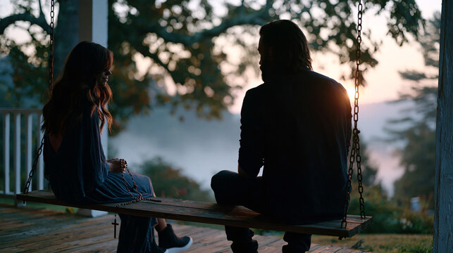 A couple sits on opposite ends of a porch swing in early morning mist, one holding a rosary and the other in quiet secular thought, a shared silence between them that communicates an ongoing and unr