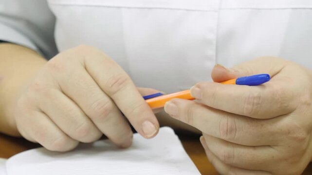 Close up shot doctor hands in white medical lab coat fidgeting with orange pen over blank notebook page on wooden desk during consultation