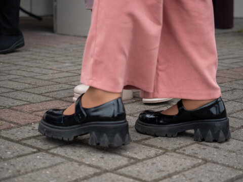 Low angle view of female legs wearing black patent leather Mary Jane shoes with thick lug soles and pink trousers standing on grey brick pavement