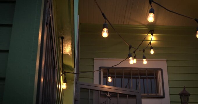 Warm bulbs hang over the doorway of a green house at dusk, casting soft light on the porch and metal gate.