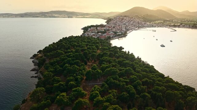 Drone flyover above a lush pine-covered peninsula toward the picturesque Greek coastal town of Ermioni glowing in warm sunset light