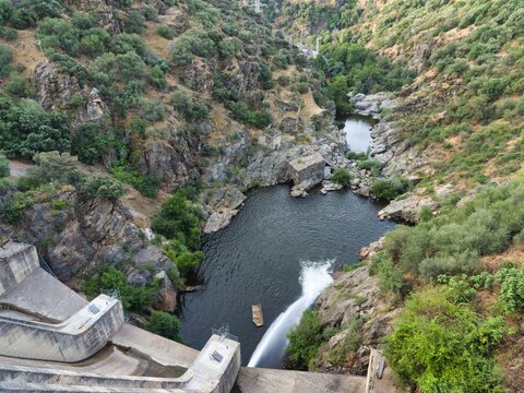 Water flows from a dam into a river surrounded by rocky hills and dense greenery in a remote area during daytime