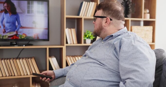 Overweight man watching television on sofa at home. He holds a remote and finds relaxation in a living room, enjoying streaming media after work. Clear concept of home media leisure lifestyle.