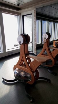 Luxury gym interior featuring elegant wooden rowing machines with water resistance near floor-to-ceiling windows. Scenic ocean view, minimalist design and functional workout equipment create an