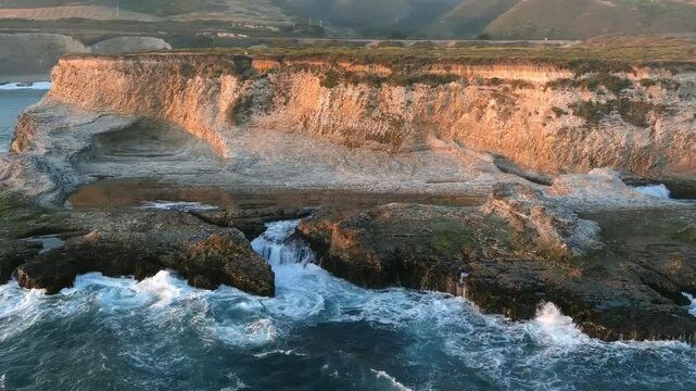Aerial flyover of waves crashing along the cliffs on the coast of California near San Francisco at sunset.