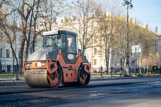 Orange road roller on street compacts freshly laid asphalt on traffic road, construction works in modern city, heavy machinery in action to pave driveway, roller smoothing surface