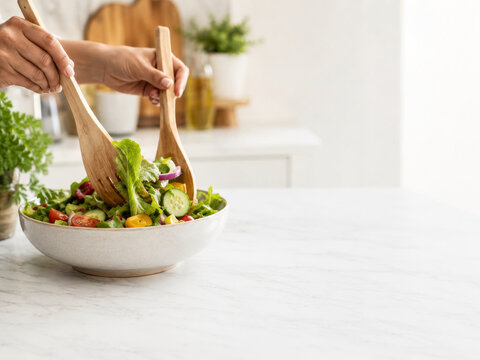 Preparaci&oacute;n de una ensalada en una cocina moderna, con manos usando cucharas de madera en un taz&oacute;n grande con verduras frescas