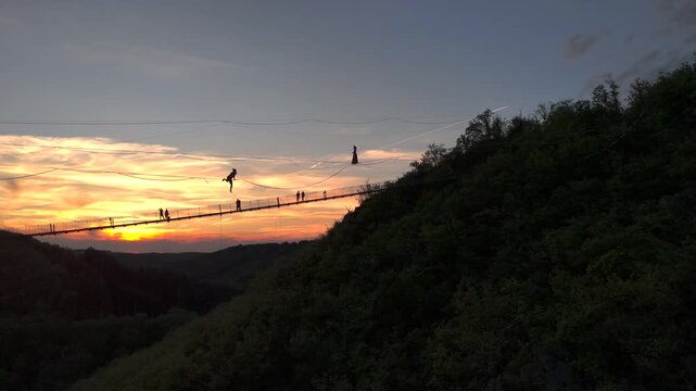 Highline Aerial Shot. The Tightrope Walker is on a rope stretched between the Mountains at high altitude at Sunset Time