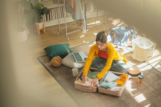 Asian woman packing clothes into open suitcase on bedroom floor surrounded by messy room, overhead view of travel preparation and trip planning lifestyle concept