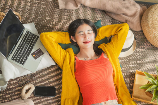 Happy Asian woman lying on floor smiling after online shopping with credit card and laptop, satisfied consumer relaxing post-purchase, fintech and lifestyle concept