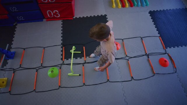 A young boy trains on an agility ladder with various obstacles in a colorful gym. The active child practices motor skills and coordination during his workout.