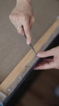 Meticulous artist carefully removing staples from the back of a canvas with a palette knife, disassembling the artwork for restoration or framing in a slow-motion, detailed close-up. vertical