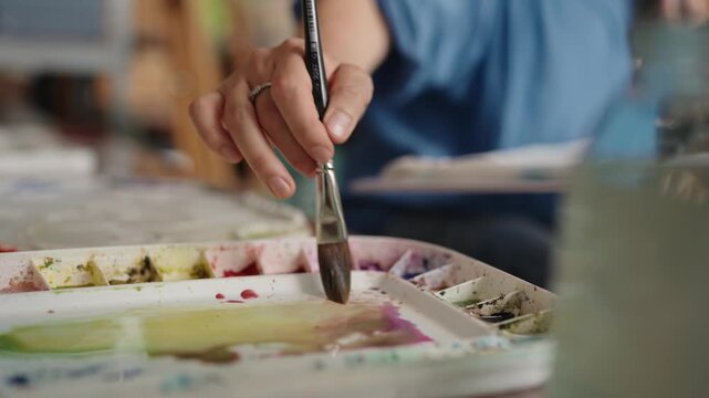 Close-up view of an artist's hand skillfully blending colorful watercolor paints with a brush on a white palette, preparing various shades for a creative artwork session in slow motion.
