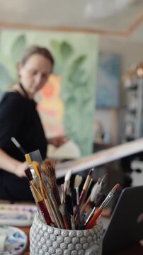Close up of a jar full of paintbrushes in an artist's studio, with the painter working on a canvas in the blurry background. Creative process in a workshop with professional art supplies. vertical