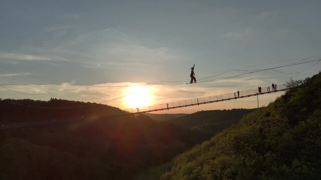 Highline Aerial Shot. Brave Girl Tightrope Walker is on a rope stretched between Mountains at high altitude, Sunset Time