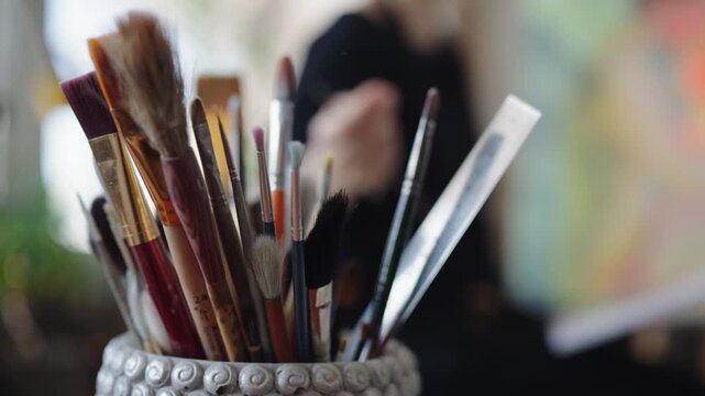 Female artist's hand selecting a paintbrush from a pot full of art supplies in a creative workshop, with a colorful canvas blurred in the background, ready for painting and inspiration.