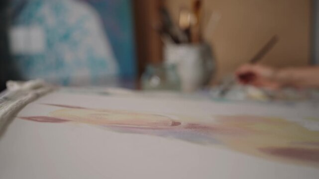 Close up view of a painter's hand using a soft brush to apply gentle pastel watercolor tones onto a white canvas, creating an abstract and artistic masterpiece in a creative workshop.