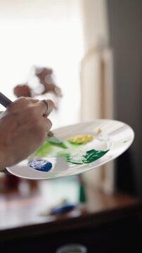 Close-up of a talented female artist's hands mixing green and yellow acrylic paint on a palette with a brush and then applying vibrant strokes to a blank canvas, creating a new masterpiece. vertical