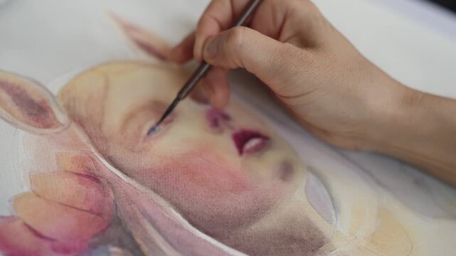Close-up of an artist's hand using a thin brush to carefully paint the eye and details of a whimsical watercolor portrait of a girl with fawn ears, highlighting the creative process