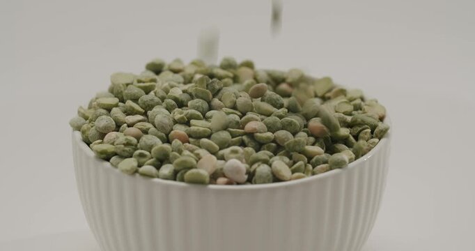 Spoon scooping and pouring raw dried green split peas into a white ceramic bowl isolated on a white background, highlighting healthy plant-based ingredients and food preparation