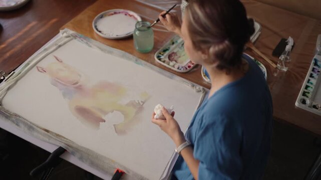 Creative woman sitting in a bright room filled with plants, painting a watercolor picture on an easel. Artistic process and inspiration in a cozy home studio with natural light