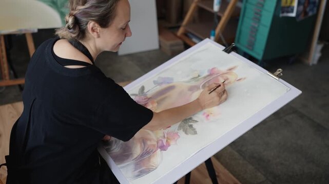 Talented female artist sitting at an easel in her creative workshop, concentrating on painting a delicate floral watercolor still life with a fine brush in a slow motion high angle view