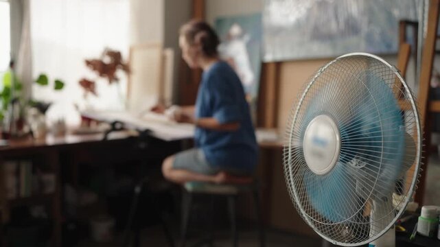 Close-up of a spinning electric fan in the foreground, cooling the air in a sunlit art studio where a blurry female painter is working on her craft, sitting at a desk by the window