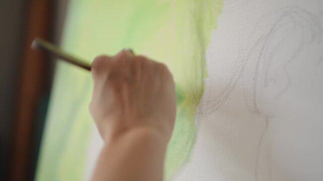 Close-up of a painter's hand using a brush to apply green paint onto a large canvas, covering a preliminary pencil sketch. The artist works with deliberate strokes in a sunlit studio space