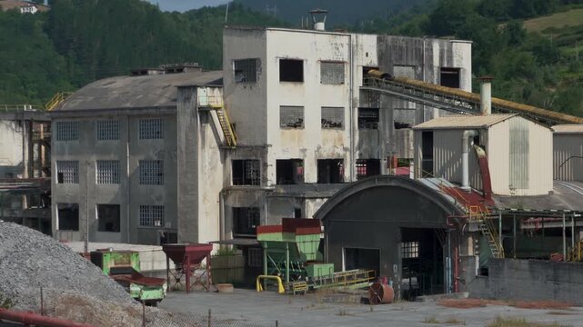 Wide view of an industrial factory complex with water tower, processing buildings, conveyors, and production facilities. Manufacturing site surrounded by green hills in northern Spain.