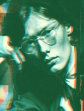 Close-up portrait of a young man wearing round glasses, shown with heavy color distortion and horizontal glitch bands on a dark background.