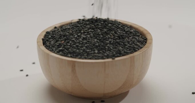 Nutritious black sesame seeds rotating in a small wooden bowl on a clean white background. Black sesame seeds falling in slow motion. Macro shot of black sesame seeds cascading down in slow motion