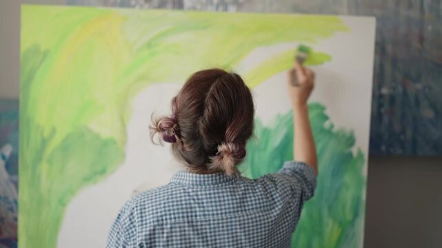 Young female artist painting with a brush on a large canvas, creating an abstract green and yellow modern masterpiece inside her messy art studio during a moment of creative inspiration