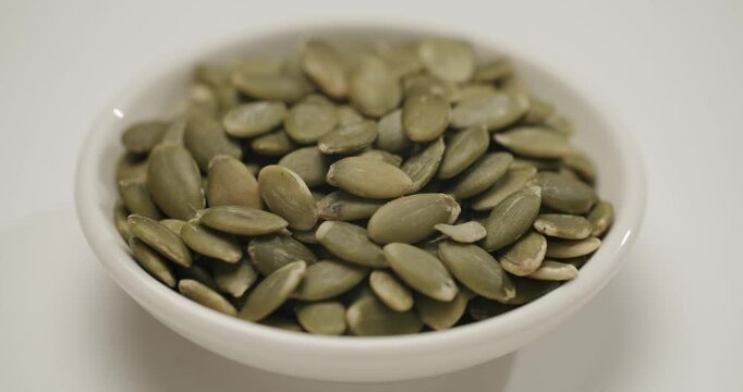 Close-up of raw, shelled pumpkin seeds, also known as pepitas, slowly rotating in a small white ceramic bowl on a white background, highlighting their nutritional and culinary value