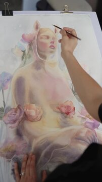 Talented female artist sitting at an easel in her creative workshop, concentrating on painting a delicate floral watercolor still life with a fine brush in a slow motion high angle view. vertical