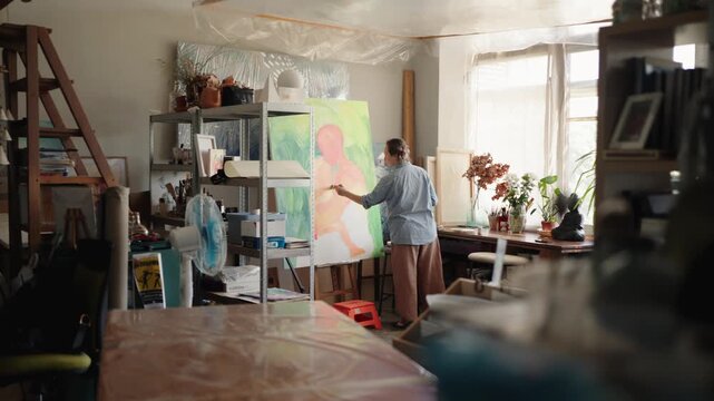 Long shot of a professional female painter creating a modern artwork on a large canvas, using a paintbrush and paints in her sunlit workshop filled with art supplies and plants