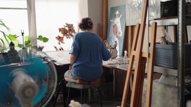 Inspired woman sitting barefoot at her desk and creating a new watercolor painting in her sunny art studio, surrounded by her artworks, brushes, and plants for a creative atmosphere