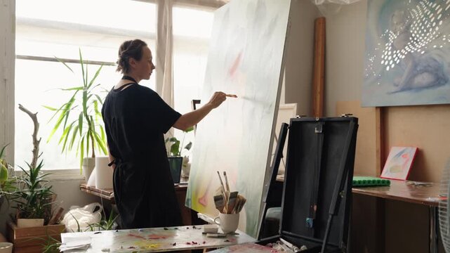 This reveals her creativity and dedication. Focused female artist with a paintbrush working on a large, abstract painting on an easel in her naturally lit art studio, colorful brushstrokes on canvas