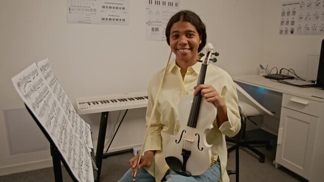 Portrait of African American teenager smiling at camera while holding violin in music classroom. Clip for promoting music education, arts programs, or online learning platforms