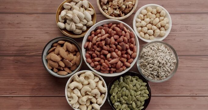 Top view of a variety of healthy nuts and seeds in bowls rotating slowly on a wooden surface, including almonds, peanuts, walnuts, cashews, pistachios, hazelnuts, pumpkin and sunflower seeds