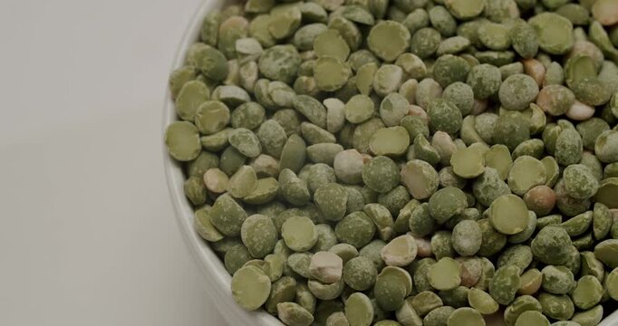 Dried green split peas in a white ceramic bowl rotating slowly. Green dried split peas rotating slowly. Spoon scooping and pouring raw dried green split peas into a white ceramic bowl