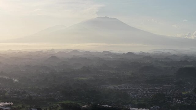 Aerial view of a suburban city at sunrise, showing rooftops, streets and green spaces with a distant volcanic mountain silhouette under soft morning light and hazy atmospheric layers